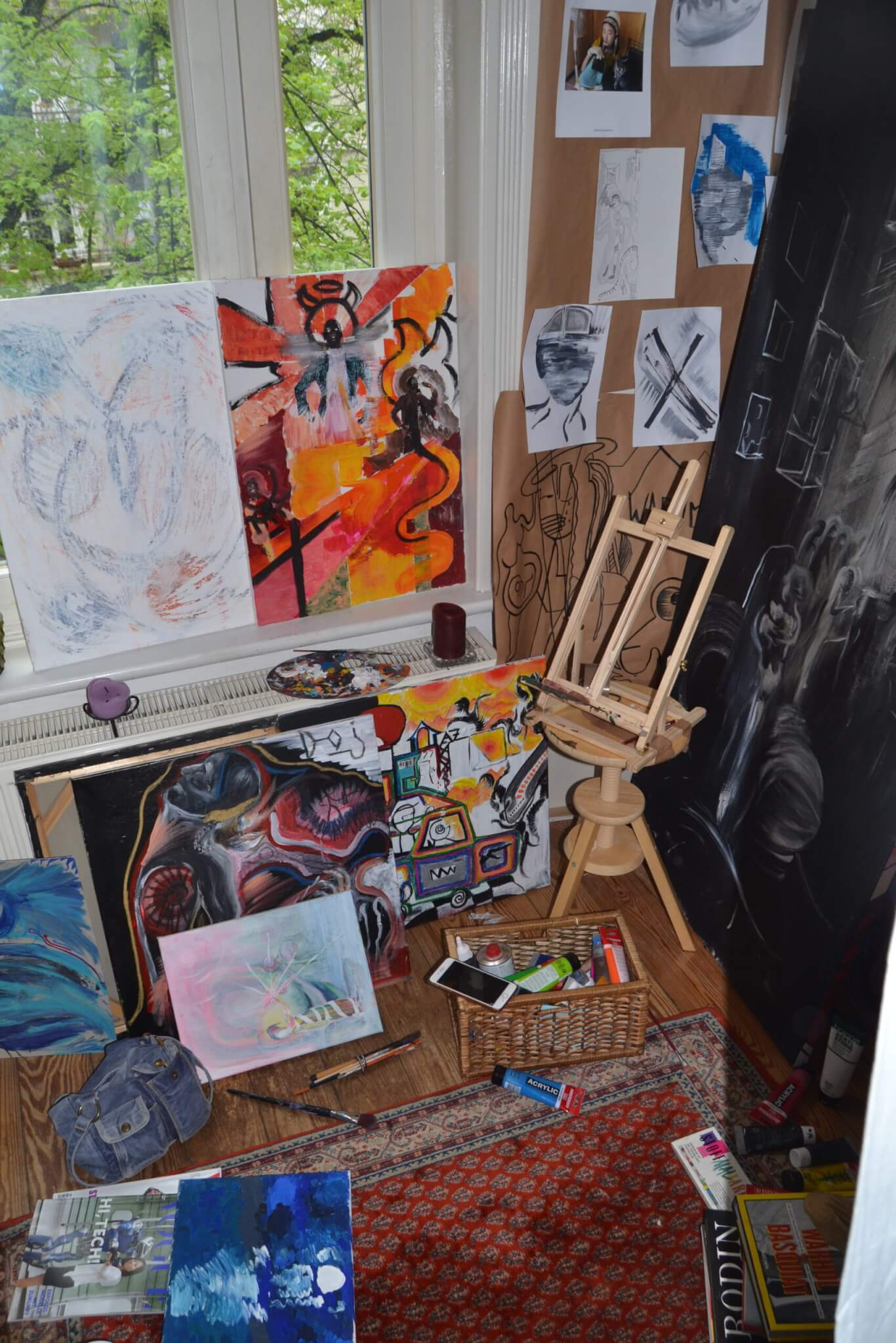 a room corner, cluttered with art supplies, drawings and paint tubes, in the background a window and trees