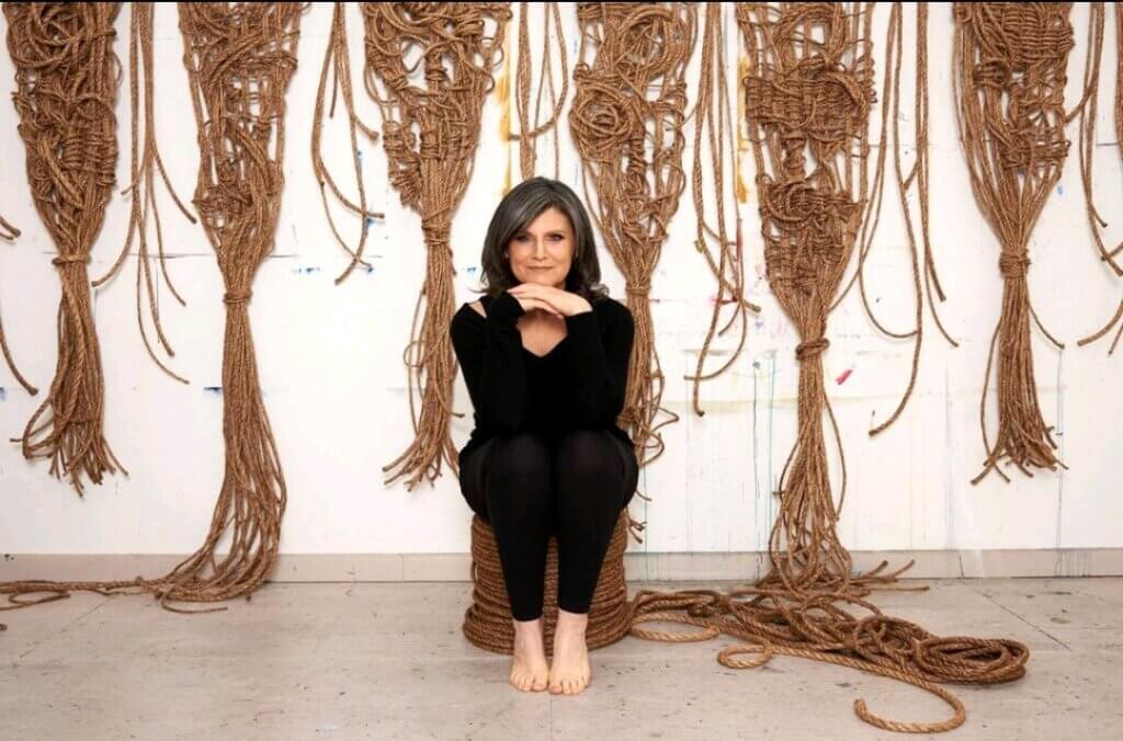 A women, Susan beallor Snyder, sits on a pile of rope, in the background are hanging six artworks of hers