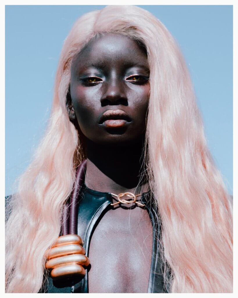 a black women with pastel pink hair and yellow contact lenses is looking directly into the camera, in the background blue sky