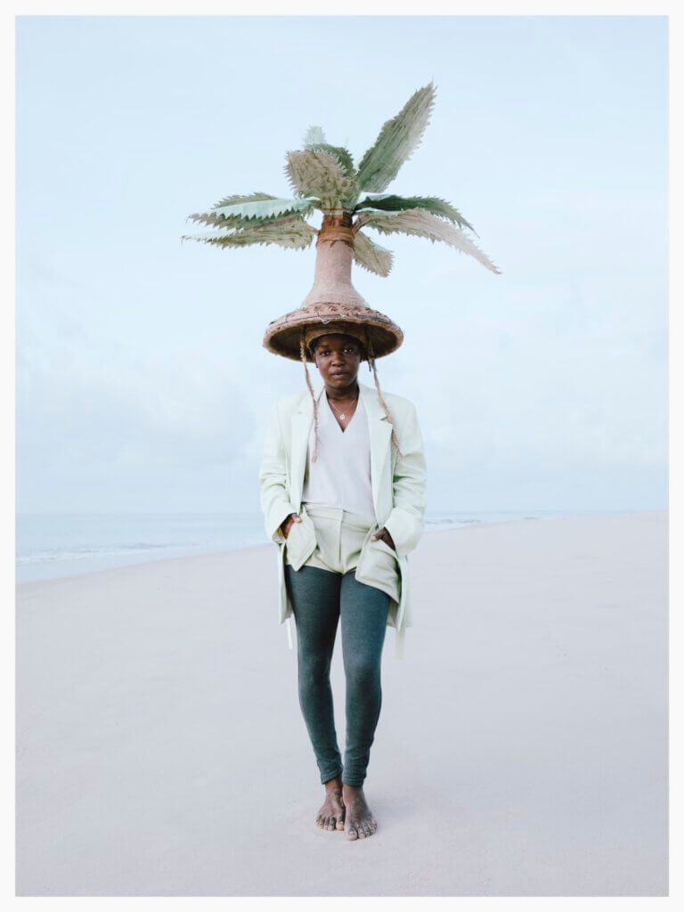 women standing on a white beach, the ocean and the sky in the background are also very light-coloured. she is waring a big head that looks like a palmtree