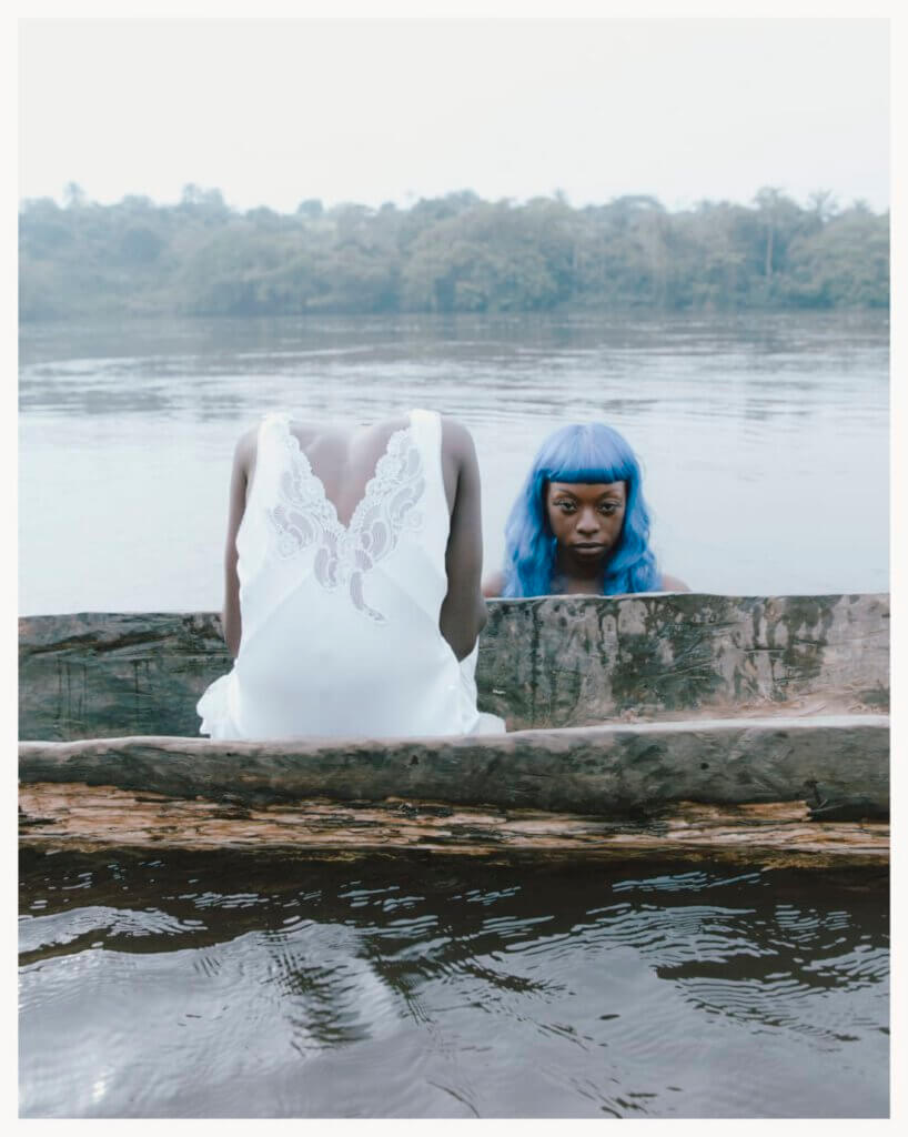 a body in a white dress sits headless on a boat. Next to it there is a womens head with long blue hair. she is looking directly into the camera