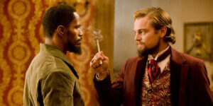 Calvin Candie, holding a cigarette holder, confronts Django in an intense standoff inside an opulent room.