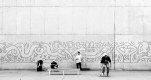 A black-and-white image of a concrete wall featuring street art in the style of Keith Haring, with people sitting and skateboarding in front of it.