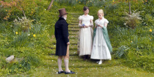 A spring garden scene from the film Emma., featuring two women in Regency-era dresses and a gentleman in a top hat conversing on a grassy path lined with daffodils.