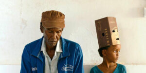 A still from The Village Next to Paradise showing a man in a brown cap and blue jacket sitting beside a young boy wearing a box with eyeholes on his head, against a bare wall.