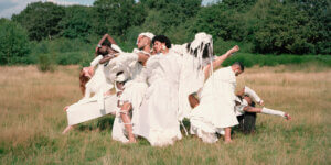 A group of diverse dancers wearing flowing white garments pose in a dynamic formation in a sunlit field, blending fashion, movement, and surrealism.