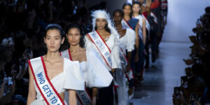 Fashion models wearing political sashes reading “Who Gets to Be American?” on a runway, highlighting immigration and labor rights in the fashion industry.