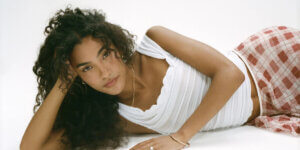 Olivia Dean lies on her side against a white backdrop, wearing a white ruched top and a red-checkered skirt, gazing softly at the camera.