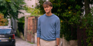 A young man with soft brown hair and a gentle smile stands on a quiet, sunlit street wearing a light blue sweater—Conrad Fisher from The Summer I Turned Pretty.