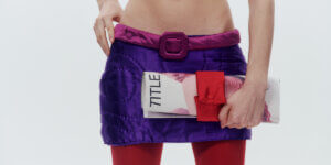 Model holding a rolled TITLE magazine secured with a red quilted Cult Form accessory, styled with a purple quilted mini skirt, pink belt, and red tights.