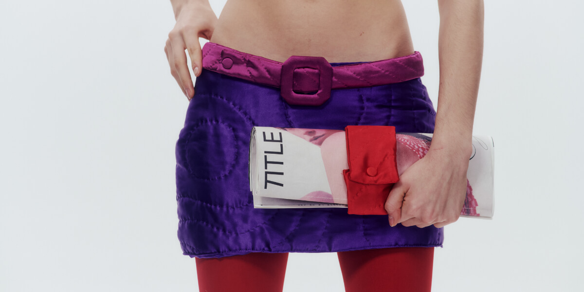 Model holding a rolled TITLE magazine secured with a red quilted Cult Form accessory, styled with a purple quilted mini skirt, pink belt, and red tights.