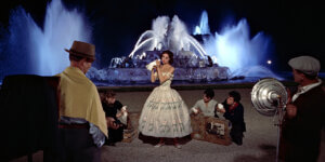 A glamorous nighttime scene from the movie Funny Face, showing a woman posing in a vintage floral dress before a dramatic illuminated fountain while a film crew surrounds her.