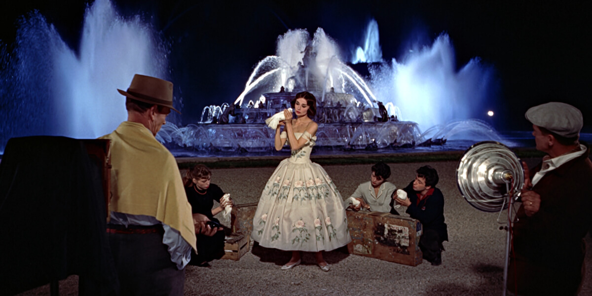 A glamorous nighttime scene from the movie Funny Face, showing a woman posing in a vintage floral dress before a dramatic illuminated fountain while a film crew surrounds her.