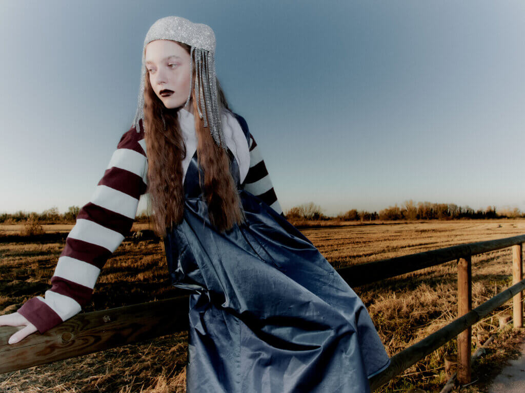 Fashion editorial set in the Italian countryside featuring a model in a soft knit look and statement accessories, styled against winter fields with a floral headpiece, photographed by Pietro Groff.