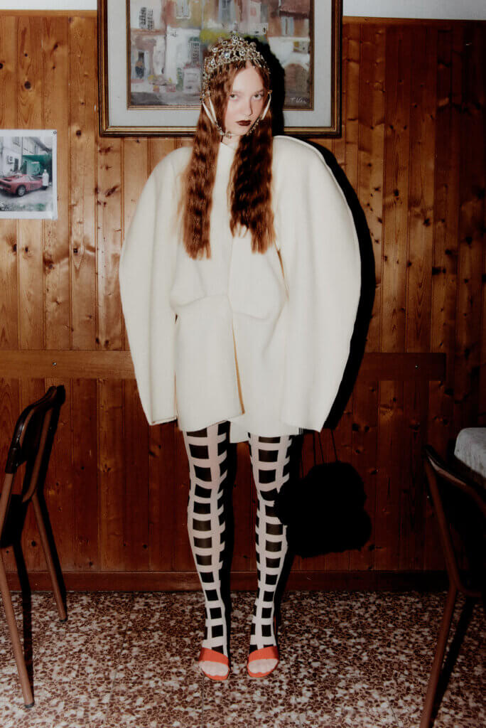 Fashion editorial set in the Italian countryside featuring a model in a soft knit look and statement accessories, styled against winter fields with a floral headpiece, photographed by Pietro Groff.