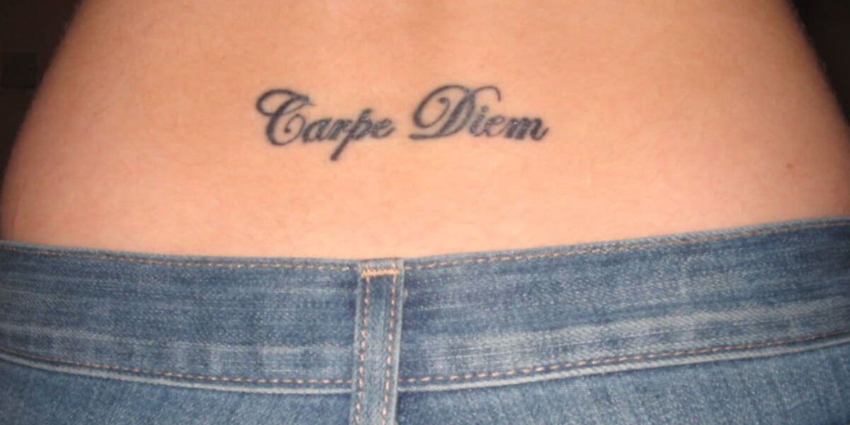 Lower back tattoo reading “Carpe Diem” above denim jeans, symbolizing living in the moment and modern YOLO philosophy.