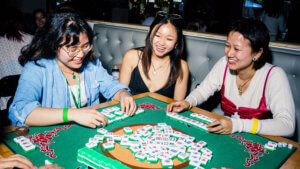 Three young Asian women playing a game of Mahjong and laughing together around a green felt table.
