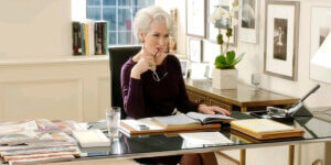 Meryl Streep as Miranda Priestly sitting at her desk in "The Devil Wears Prada," holding glasses to her mouth while looking at a book.