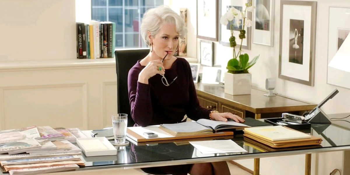 Meryl Streep as Miranda Priestly sitting at her desk in "The Devil Wears Prada," holding glasses to her mouth while looking at a book.