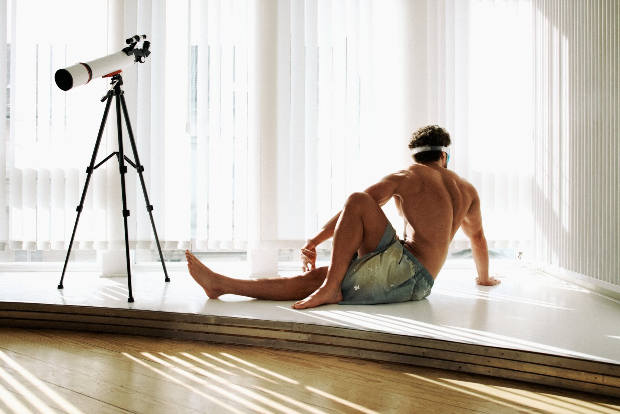 Arda Saatçi stretches over his knee on the floor wearing blue shorts in front of drawn window shades with a telescope beside him