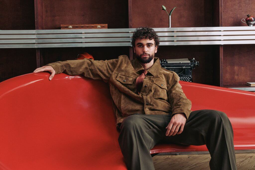 Arda Saatçi sits on a modern red couch in a brown suede blazer, green tie and dark brown pants