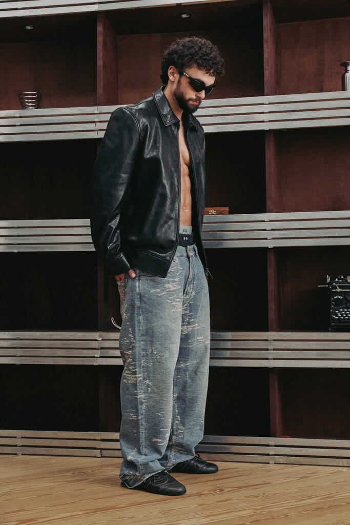 Arda Saatçi shirtless in a leather jacket, sunglasses, jeans and black shoes standing indoors in front of a metal shelf