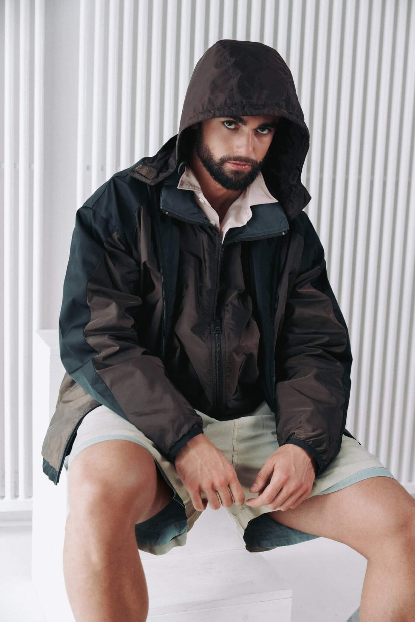 Arda Saatçi sits with his hood up wearing a brown jacket and shorts for TITLE Magazine’s Berlin Psycho fashion editorial
