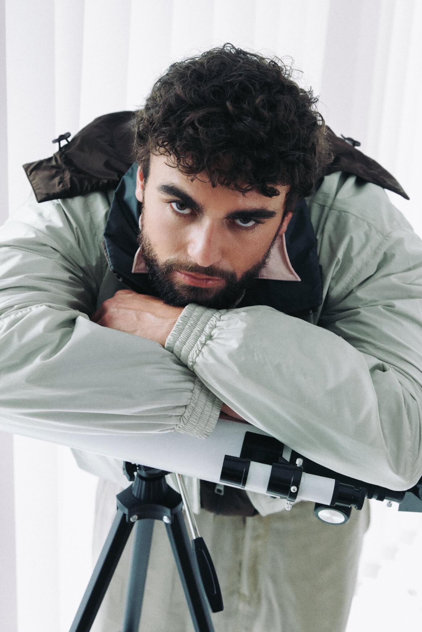 Arda Saatçi leans on a telescope wearing a gray and brown jacket in TITLE Magazine’s Berlin Psycho fashion editorial