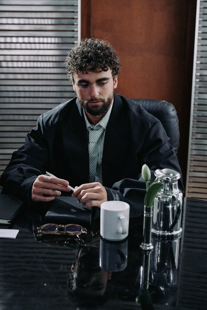 Arda Saatçi sits at a desk in a suit and tie holding a pen for TITLE Magazine’s Berlin Psycho fashion editorial