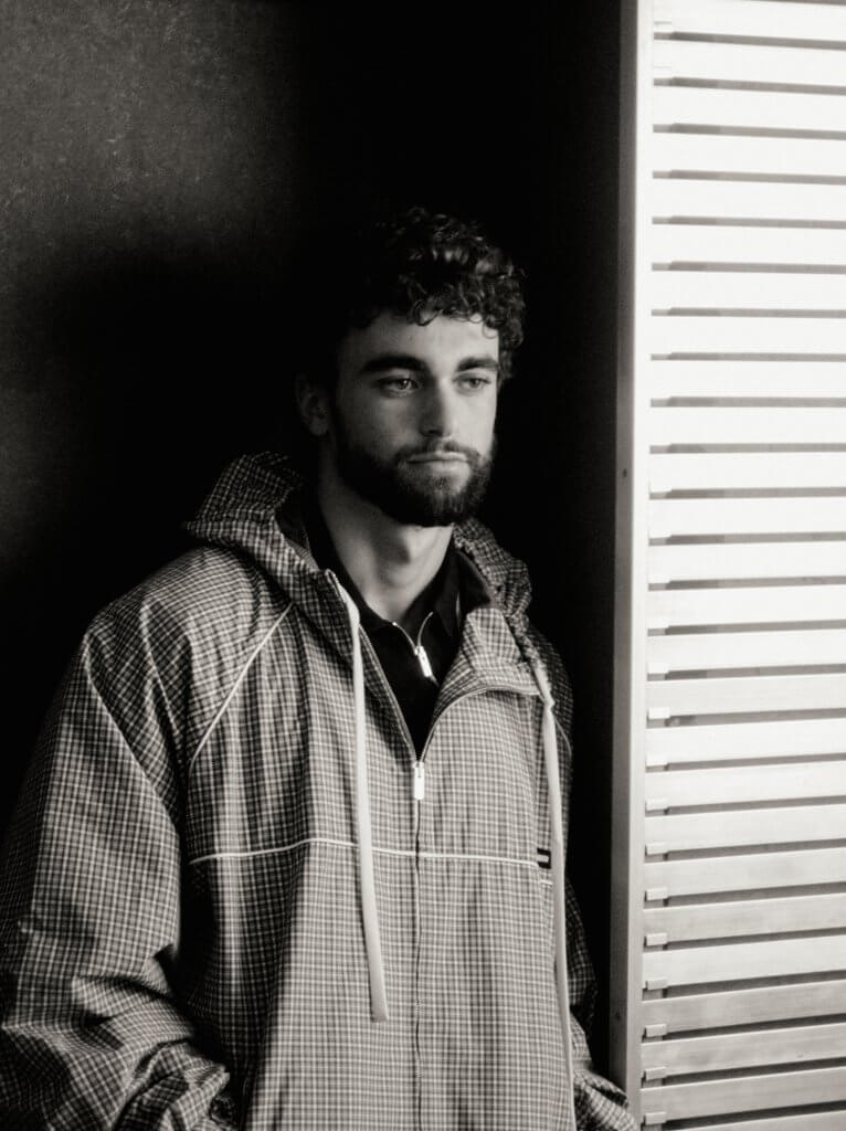 Arda Saatçi stands indoors wearing a checkered windbreaker and jeans for TITLE Magazine’s Berlin Psycho fashion editorial