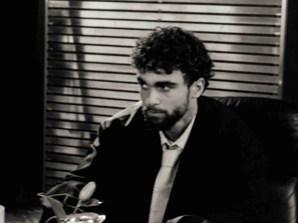 Arda Saatçi sits at a desk in a suit and in black and white for TITLE Magazine’s Berlin Psycho fashion editorial