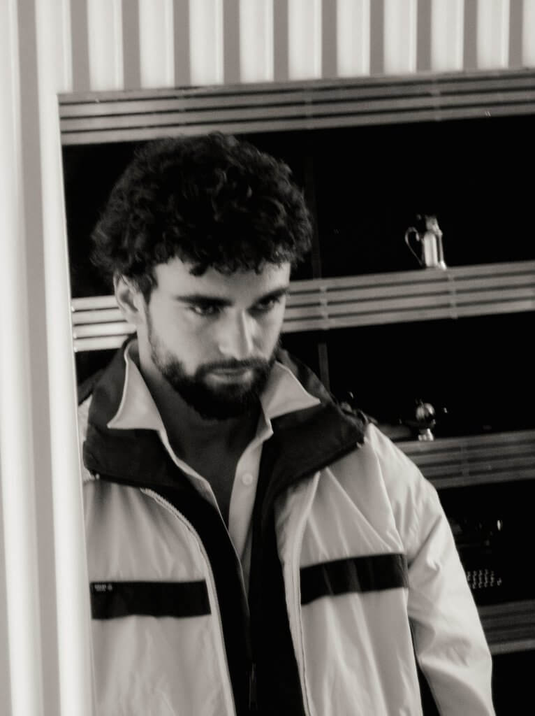 Arda Saatçi stares at his reflection in a mirror wearing a light windbreaker in a black-and-white fashion editorial