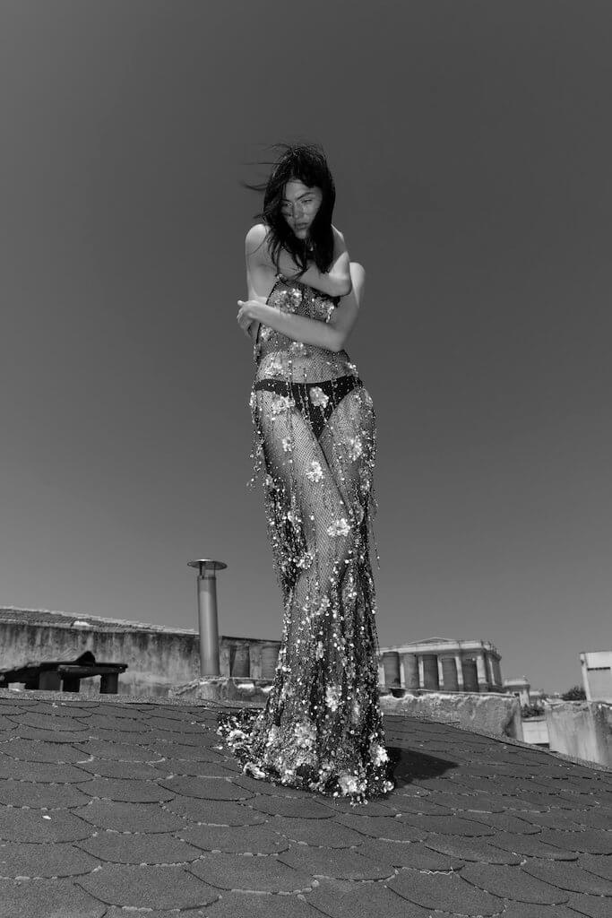 Full-length black-and-white fashion photograph of a model in a sparkling sheer floral gown standing barefoot on a rooftop, holding herself against the wind with her arms crossed.